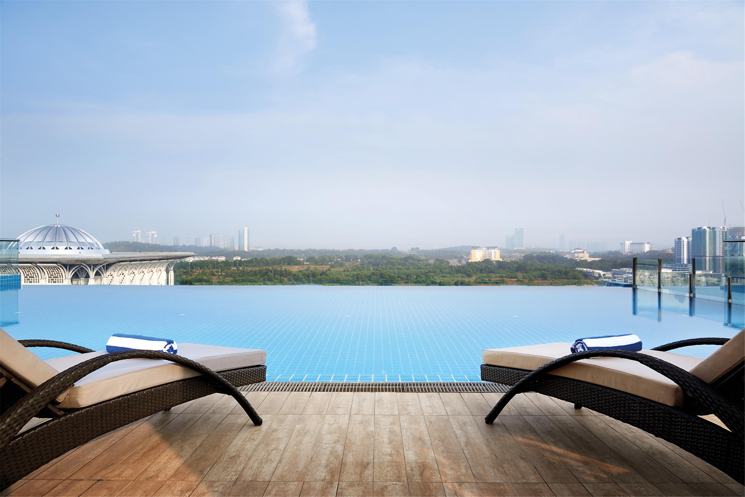 Отель Dorsett Путраджая Экстерьер фото The photo depicts a luxurious outdoor space featuring an infinity pool that extends toward the horizon, blending seamlessly with the sky. In the foreground, there are two sun loungers with rolled towels on each, positioned on a wooden deck. The backd