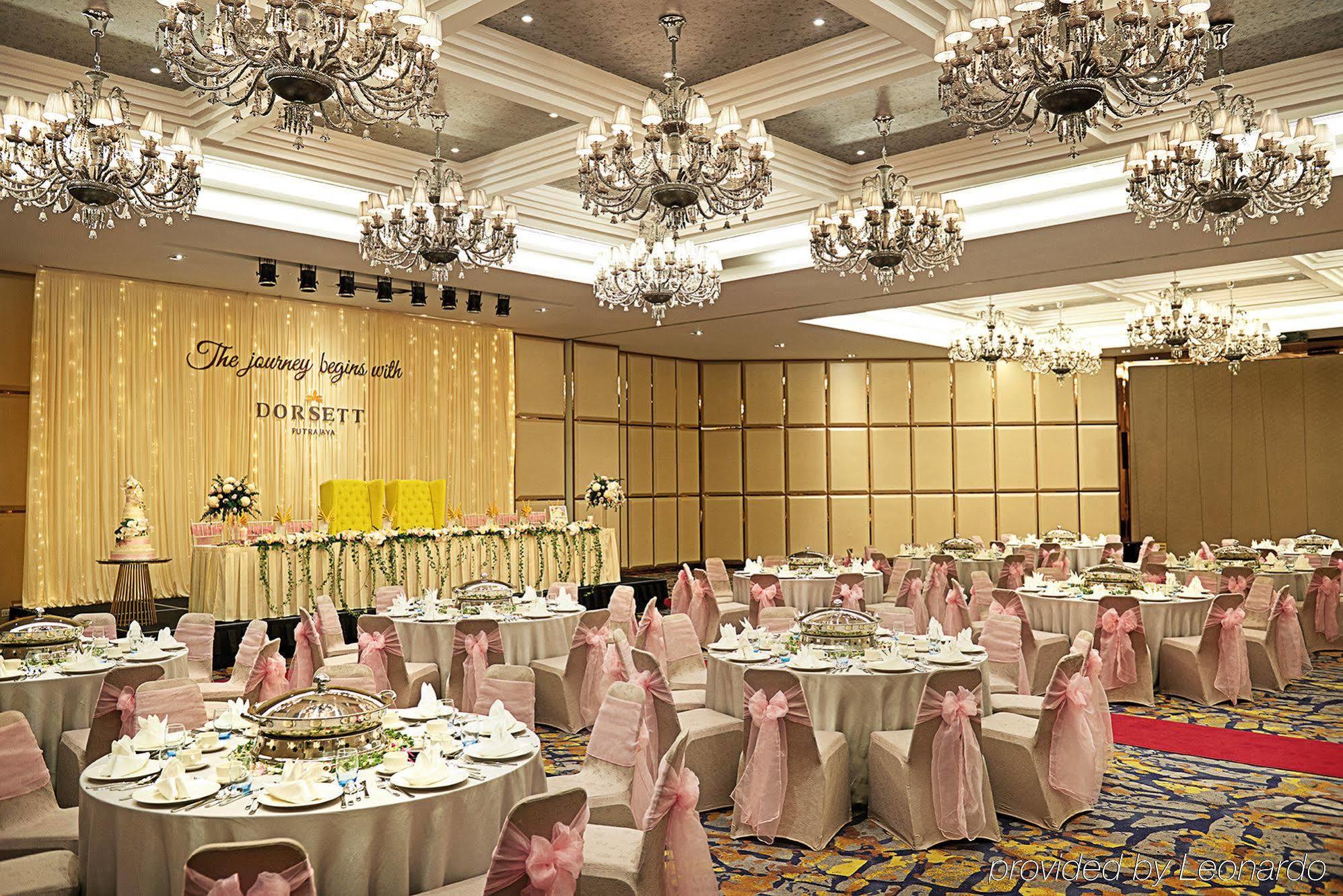 Отель Dorsett Путраджая Экстерьер фото The photo shows an elegant banquet hall set up for a celebration or event. The room features numerous round tables covered with white tablecloths and adorned with pink sashes on the chairs. There are decorative centerpieces on the tables, and the wal