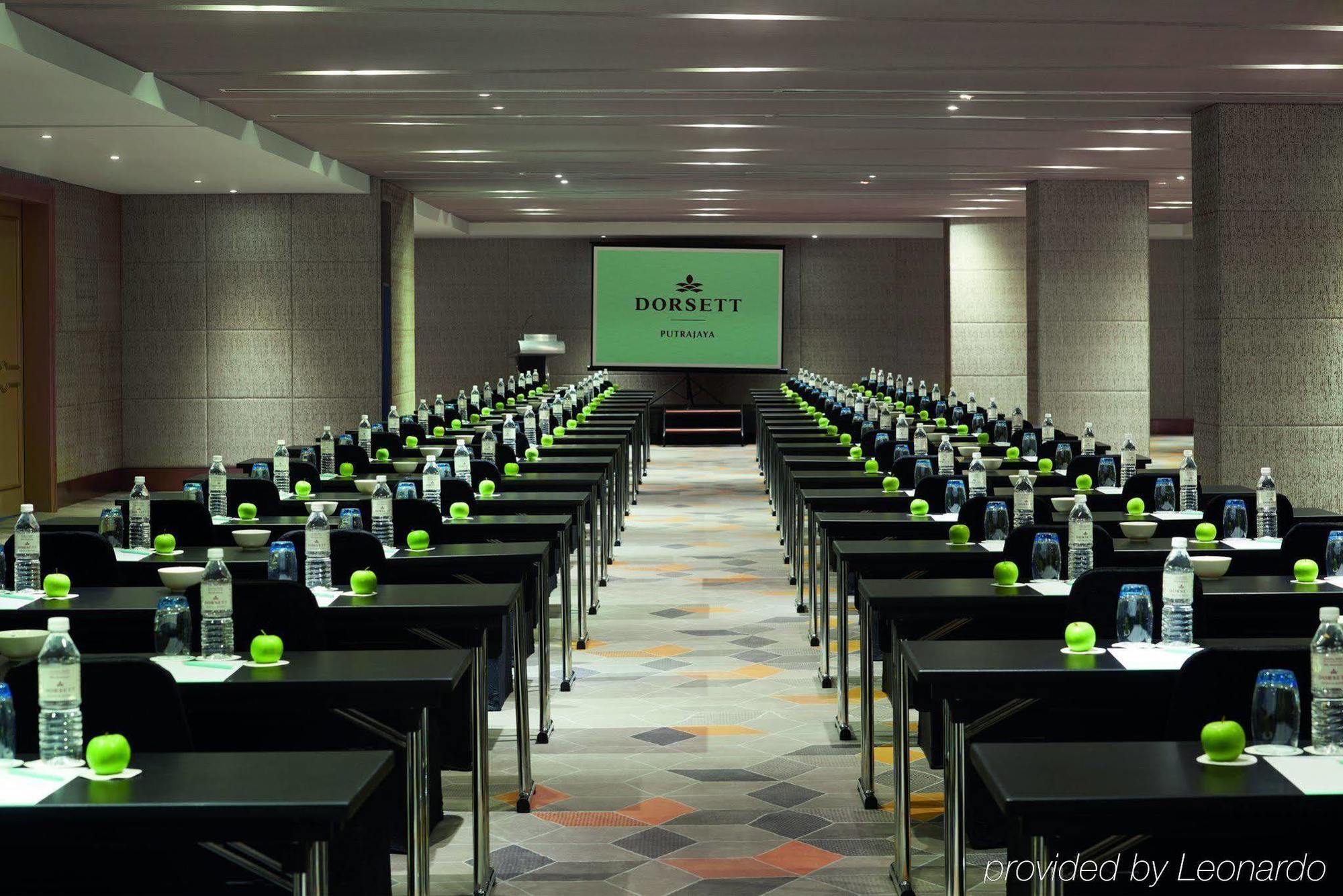 Отель Dorsett Путраджая Экстерьер фото The photo shows a conference room set up for a meeting or event. There are multiple rows of black tables arranged neatly, each equipped with a green object, possibly a plant or decorative item, and a bottle of water. In the center of the room, there 