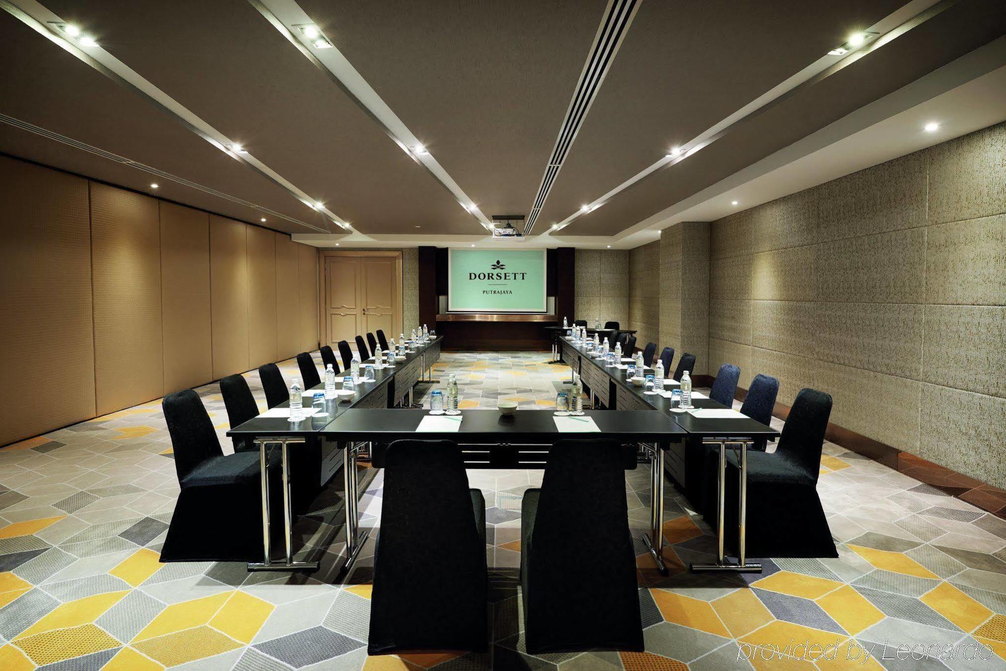 Отель Dorsett Путраджая Экстерьер фото The photo shows a modern conference room arranged for a meeting. In the center, there is a large rectangular table surrounded by sleek black chairs. At the front of the room, there's a presentation screen with the name or logo displayed. The walls ar