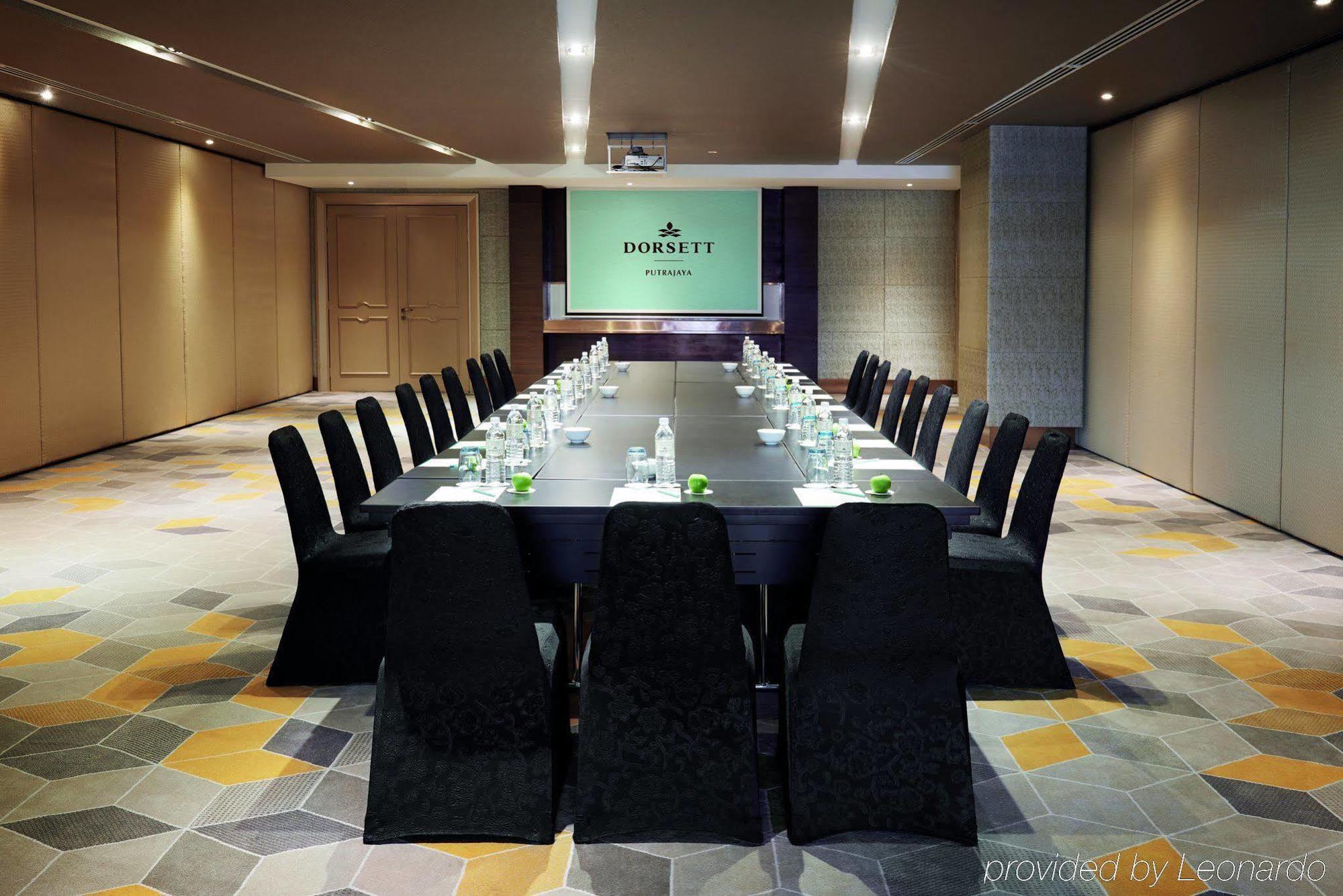 Отель Dorsett Путраджая Экстерьер фото The photo shows a modern conference room set up for a meeting. A long, dark wooden table is surrounded by black chairs with a sleek design. On the table, there are bottles of water and small green plants or decorative items. At the head of the room, 