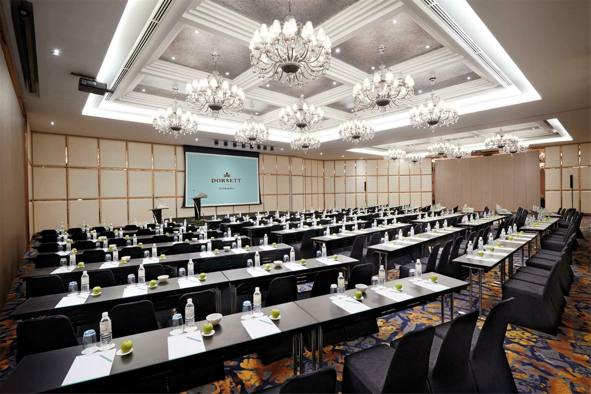 Отель Dorsett Путраджая Экстерьер фото The photo shows a large conference room set up for an event. It features a number of black tables arranged in rows, each equipped with water bottles and small green apples. The room has a modern and elegant decor, highlighted by ornate chandeliers ha